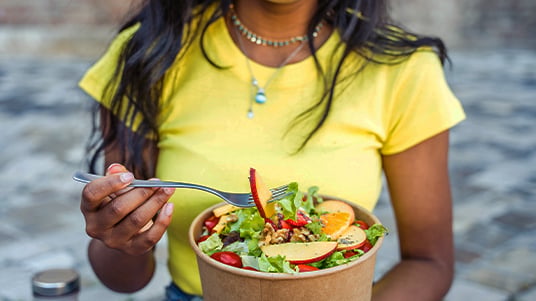 Close-up of a person holding a fork above a bowl of fresh salad with leafy greens, sliced vegetables, and colorful toppings, seated outdoors in a casual dining setting.