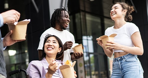 Group of people standing outdoors near a modern building, holding takeout food boxes and utensils while socializing in a bright, urban setting.