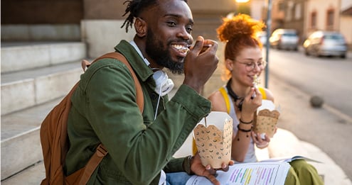 Two people sitting on outdoor steps, eating from takeout food containers, with backpacks and printed papers beside them in a casual urban setting.