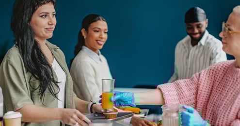 People standing indoors at a food service counter, receiving meals in cardboard containers from staff wearing gloves, with a modern interior and blue wall in the background.