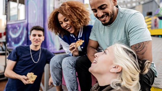 Group of people sitting outdoors near colorful structures, sharing slices of pizza and chatting together in a casual urban setting with food trucks in the background.
