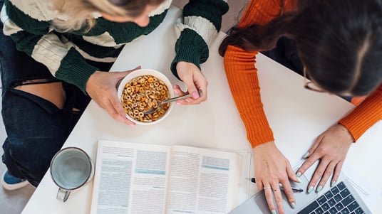 Top-down view of people studying at a table with an open book, laptop, coffee mug, and a bowl of cereal, combining coursework and casual dining in a shared workspace.
