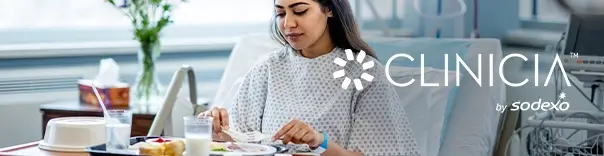 Person in a hospital bed wearing a gown, eating a meal from a tray with food and drinks; medical equipment and a sunlit window in the background.