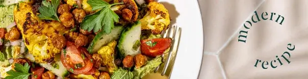 Plate of food with roasted cauliflower, chickpeas, sliced cucumbers, cherry tomatoes, and fresh parsley garnish; a fork is visible on the right side of the plate.