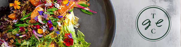Vibrant vegetable salad with edible flowers on a dark plate, with circular Tgec logo on the right.