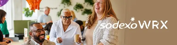 Group of people in a social or professional setting, possibly a cafeteria or break room, with balloons and plants in the background.