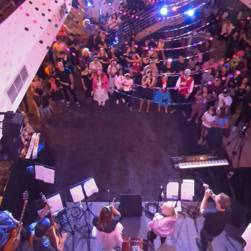 A party with pink lighting, streamers slung around the ceiling, a band is playing and there's a crowd on the dance floor.