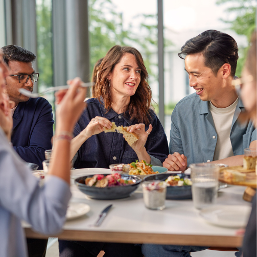 A group of diverse individuals are sitting, eating and laughing around a table.