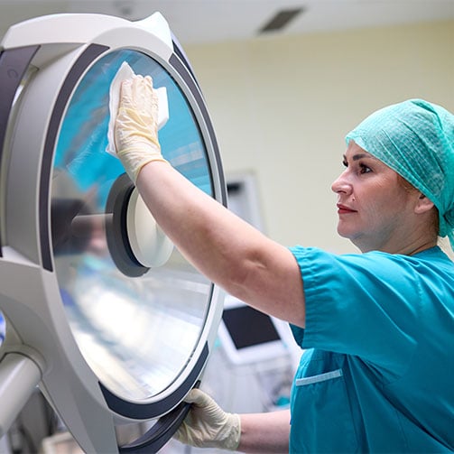 Krankenhausmitarbeiterin in OP-Kleidung reinigt sorgfaeltig eine Operationsleuchte im OP-Saal.