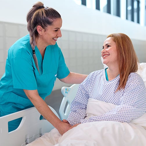 Freundliche Pflegekraft haelt die Hand einer laechelnden Patientin im Krankenhausbett.