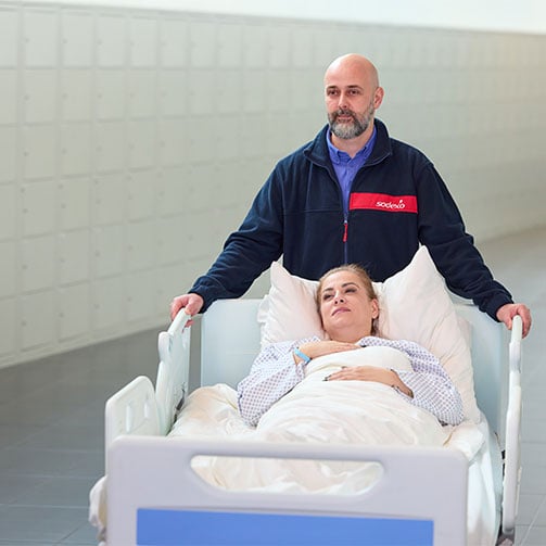 Fahrtendienstmitarbeiter schiebt eine Patientin im Krankenbett durch einen hellen Klinikflur.