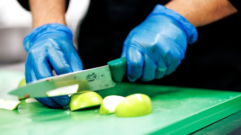 Koch mit Hygiene- Handschuhen schneidet praezise gruenen Apfel mit scharfem Messer