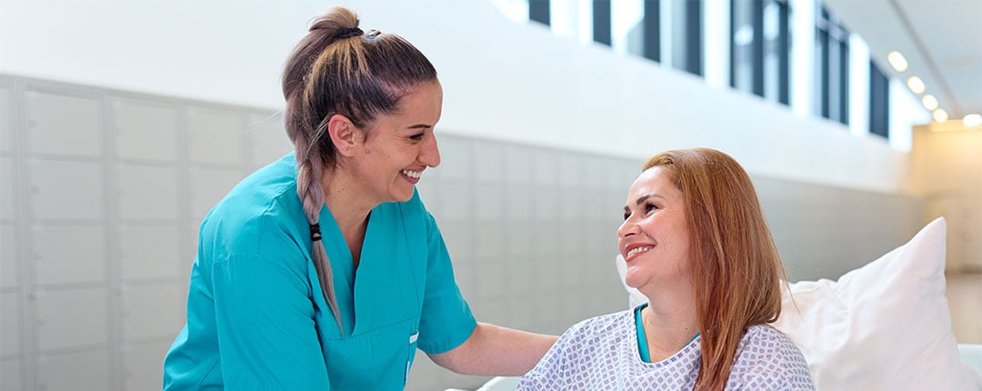 Freundliche Pflegekraft haelt die Hand einer laechelnden Patientin im Krankenhausbett.
