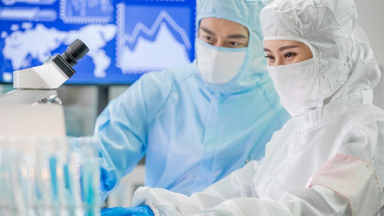 Laborsituation, eine Frau und ein Mann in Schutzkleidung und Maske stehen vor einem Mikroskop
