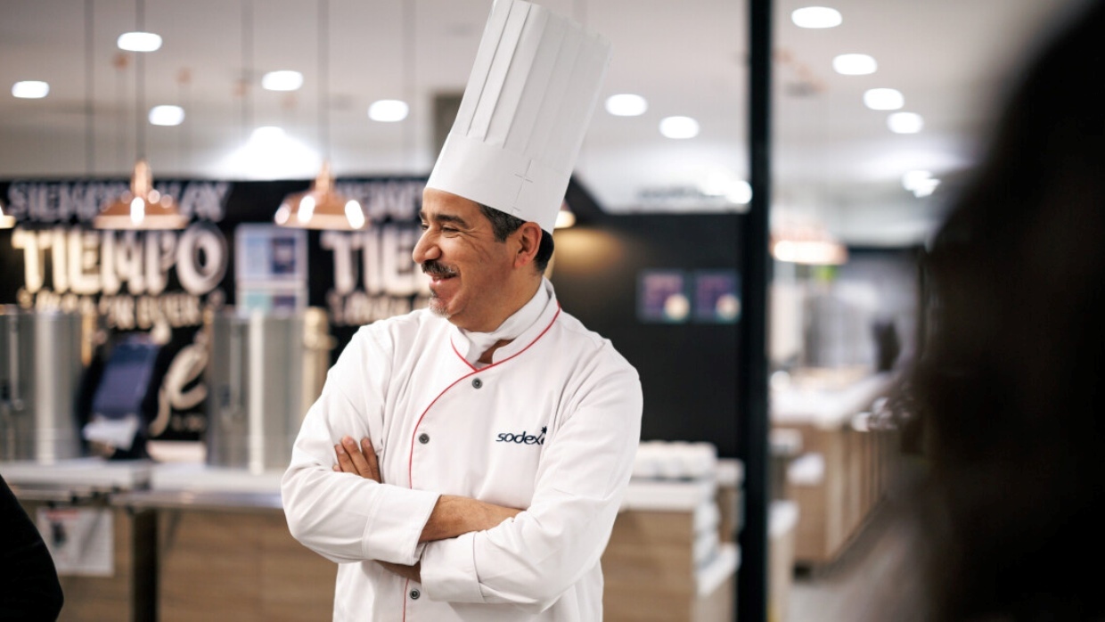 Sodexo Koch in Sodexo Uniform verschränkt seine Hände und schaut auf die linke Seite und steht in einem Sodexo Unternehmensgastronomie Restaurant