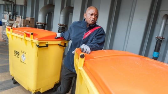 Sodexo Mitarbeitender mit oranger Weste schiebt zwei orange Abfallcontainer