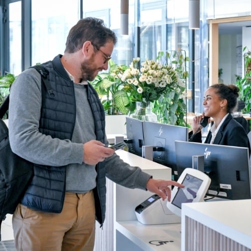 Ein Mann der als Gast zu Sodexo kommt batched sich ein und eine Sodexo Mitarbeiterin ist am Empfang am Telefon
