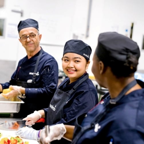 Drei Sodexo Köche sind in ihrer Sodexo Uniform in der Küche und bereiten Gemüse vor