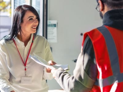Eine Sodexo Mitarbeiterin mit einem roten Lanyard gibt Unterlagen einem Mitarbeitenden mit einer roten Weste
