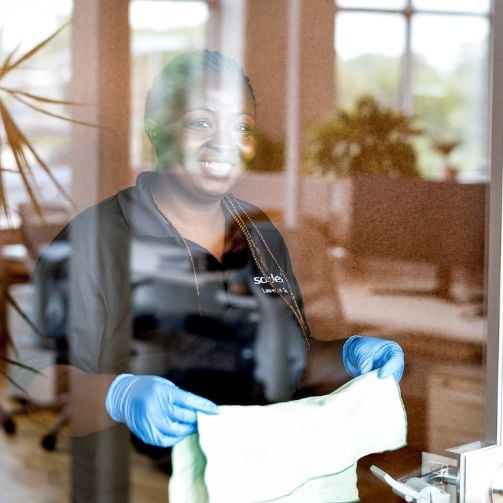 Sodexo Mitarbeitende in ihrer Uniform mit blauen Reinigungshandschuhen und einem Lappen in der Hand reinigt die Glasscheibe.