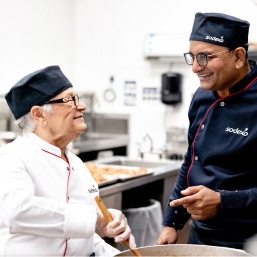 Zwei Sodexo Mitarbeitende verschiedener Generationen in Uniformen arbeiten gemeinsam in der Küche, lachen zusammen und haben Spass.