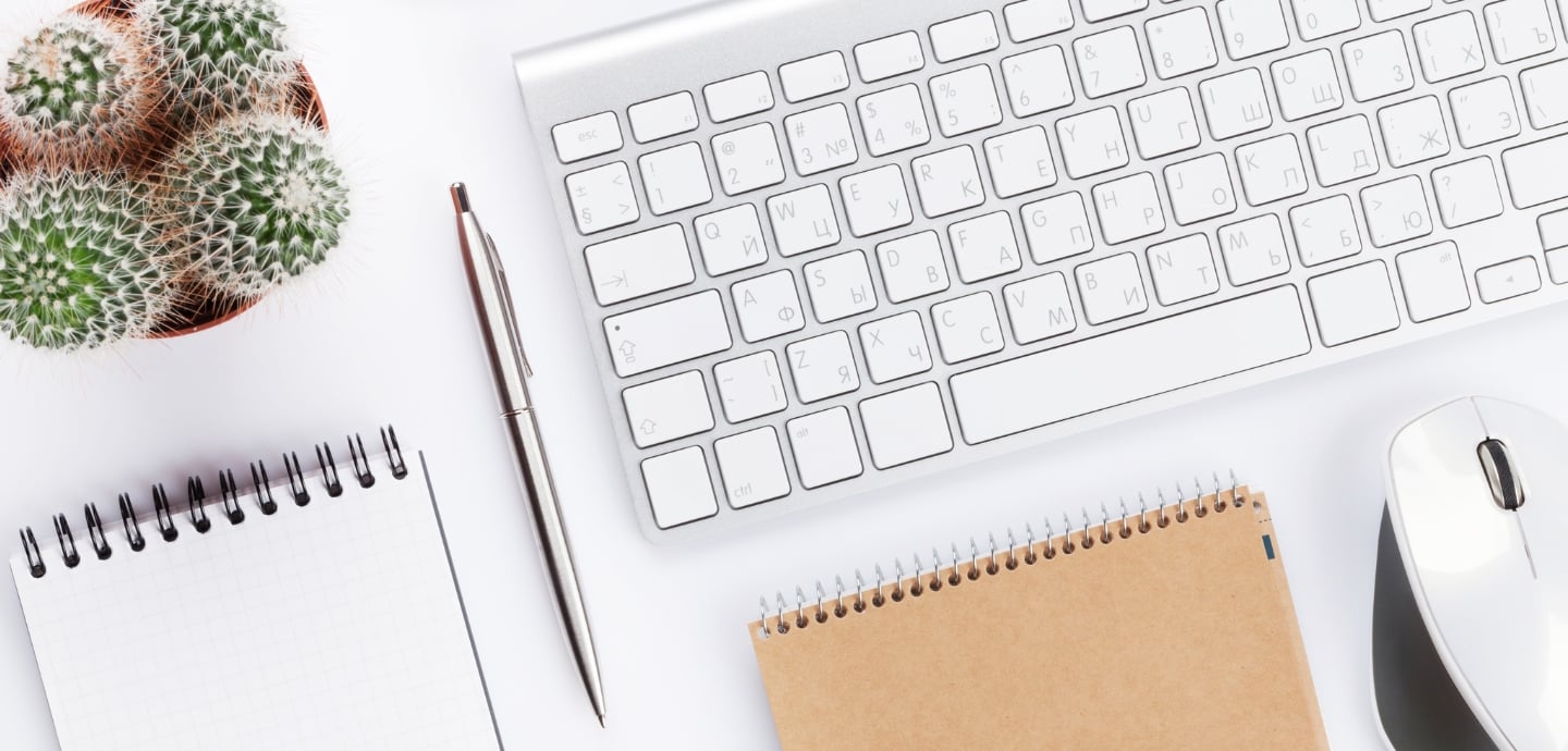 Auf einen Tisch liegt ein Kugelschreiber, eine Maus, Tastatur, zwei Bloecke und eine Kaktus