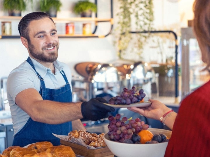 Sodexo Mitarbeiter mit blauer Schürze der hinter der Theke steht serviert einer Dame mit rotem Pulli Trauben in einem Unternehmensgastronomie Betrieb