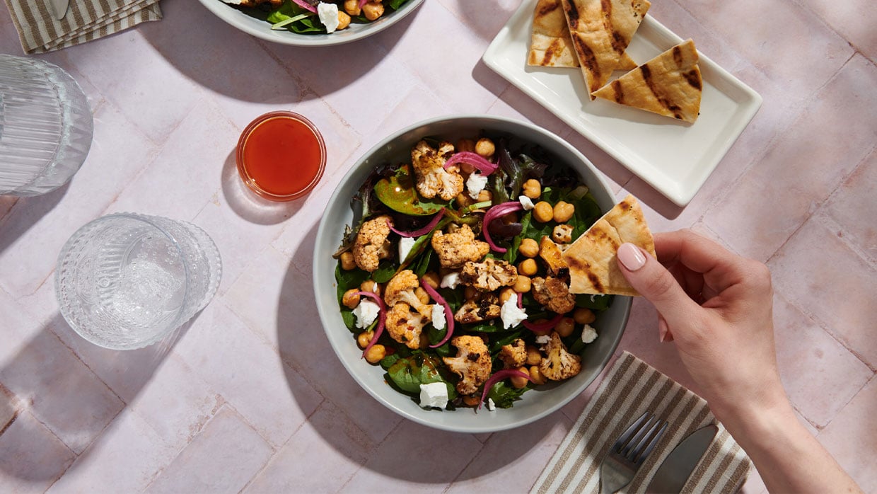 Eine pflanzenbasierte Bowl mit Gemüse, Feta und knusprigen Pitastücken. Eine Hand hält ein Stück Pita bereit zum Essen.