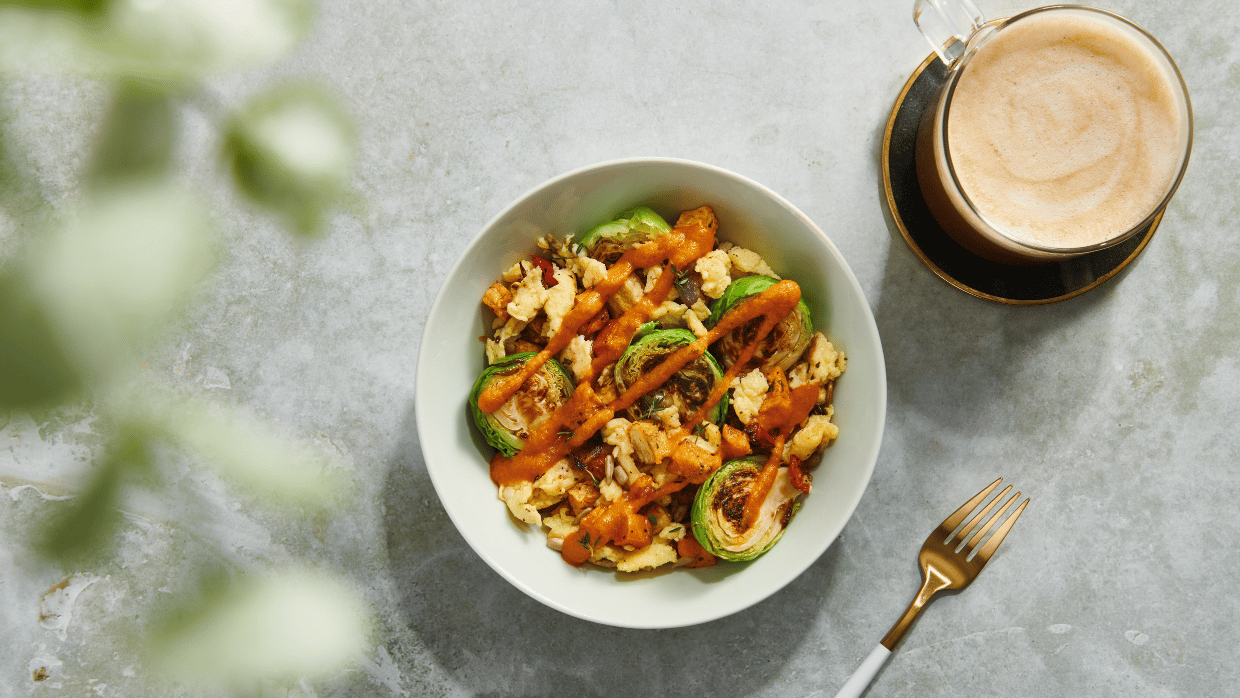 Blick von oben auf eine Gemüsebowl mit Rosenkohl und Blumenkohl auf einem grauen Tisch. Daneben liegt eine Kabel und darüber steht eine Tasse mit Kaffee.