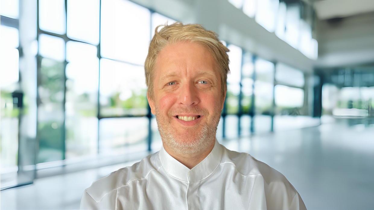 Portrait von Reuven Schockner, Head of Operational and Culinary Excellence bei Sodexo Deutschland in Kochjacke vor einer Fensterfront.