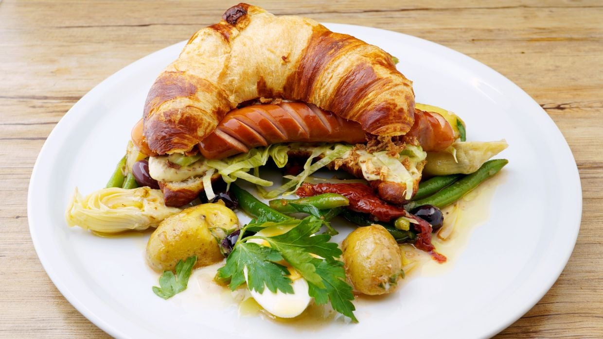 Fusion-Küche WM-Aktion: US-Hot Dog trifft französisches Croissant mit Gemüse als kreative Kombination auf einem Teller