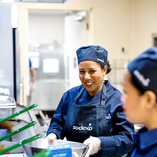 Zwei Mitarbeiterinnen in blauen Sodexo-Uniformen bereiten in einer Großküche lächelnd Gerichte für die Mitarbeiter zu.