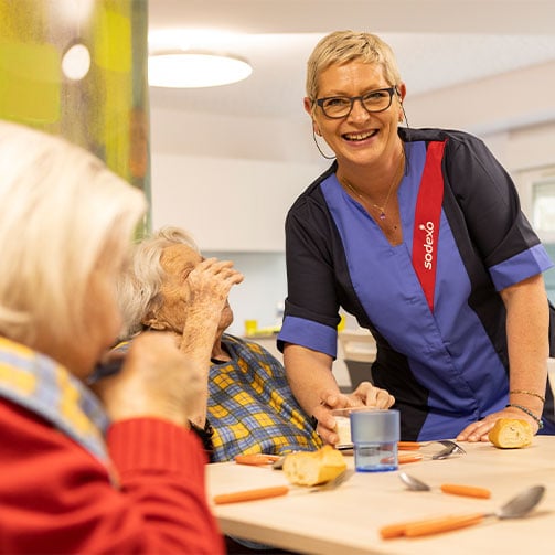 Eine Sodexo-Mitarbeiterin in einer Senioreneinrichtung, umsorgt Bewohner lächelnd mit leckerer Mahlzeit.