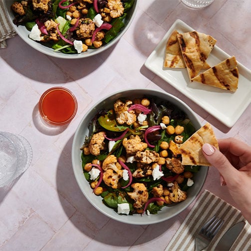 Eine pflanzenbasierte Bowl mit Gemüse, Feta und knusprigen Pitastücken. Eine Hand hält ein Stück Pita bereit zum Essen.