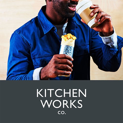 Mann in einem blauen Hemd genießt einen leckeren Wrap und einen Becher Kaffee. Das Kitchen Works Logo ist prominent im Bild zu sehen.