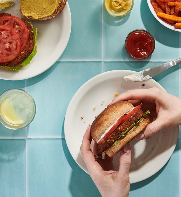 Eine Hand hält einen Burger über einem Teller, der appetitlich angerichtet ist.