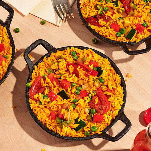 Drei große Pfannen, mit farbenfroher veganer Paella, stehen auf einem Holztisch