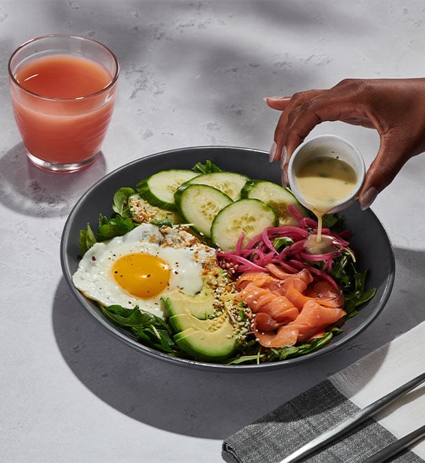 Eine Hand träufelt ein Dressing über eine farbenfrohe Salat-Bowl mit Spiegelei und knackigem Gemüse. Modernes Ambiente.