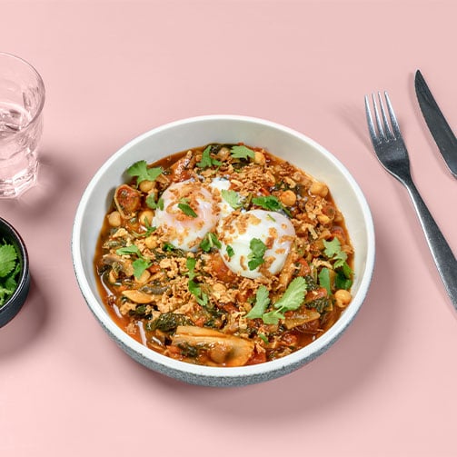 Eine Teller voller roter Shakshuka, gekrönt von zwei perfekt pochierten Eiern serviert in moderner Umgebung.