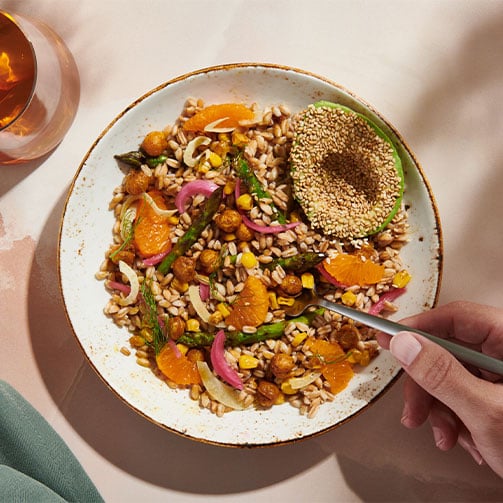 Eine bunte Gemüsebowl mit einer Avocado, bestreut mit Sesam. Eine Hand führt eine Gabel in das Gericht. 