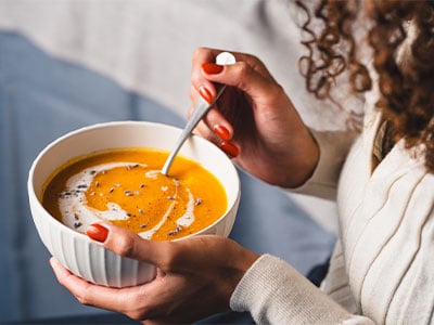 Frau mit lockigen Haaren löffelt eine Kürbissuppe aus einem weißen Teller.