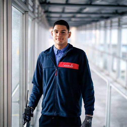 Ein Sodexo-Mitarbeiter mit dunkelblauem Fleecepulli und einem Walkie-Talkie in der Hand steht im Flur eines Firmengebäudes.