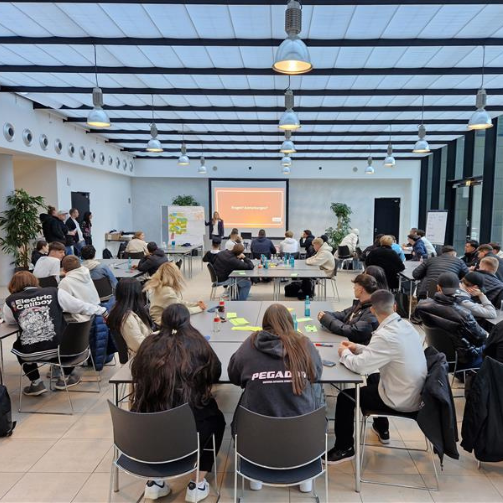 Alina Beck, Specialist People Development bei Sodexo Deutschland, stellt den Auszubildenden in einer Präsentation das Unternehmen vor. Die Auszubildenden sitzen an Gruppentischen und hören zu.