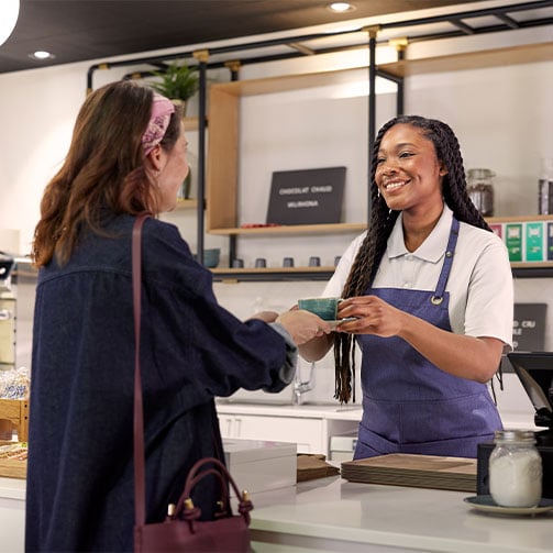 Eine Sodexo-Mitarbeiterin in Schürze reicht einer anderen Frau eine Tasse Kaffee in einer modernen Kantine