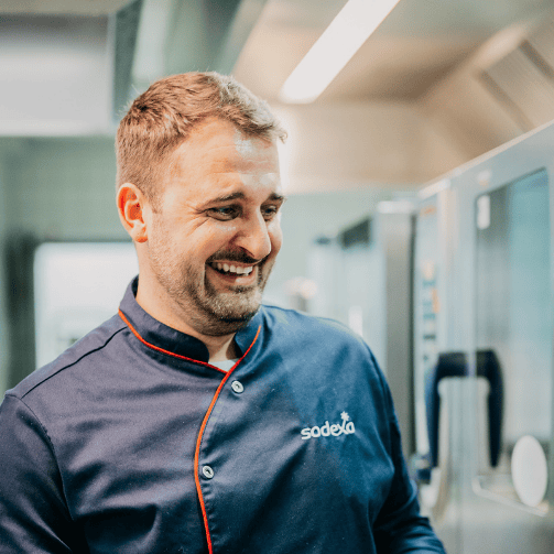 Sodexo Food-Experte Jascha Händle in Nahaufnahme lachend in Kochuniform in einer Großküche.