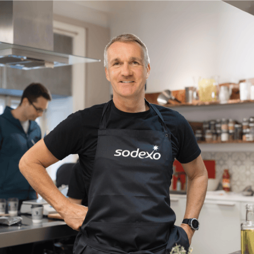 Food-Experte und Rezepte-Entwickler Thomas Hopp von Sodexo posiert in Sodexo-Schürze in einer Küche für die Kamera.