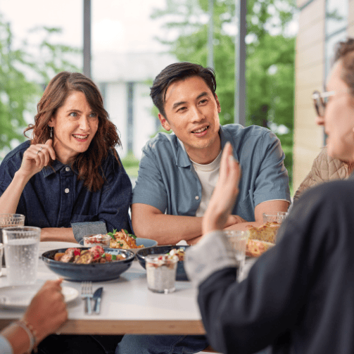 Vier Gäste sitzen an einem Tisch mit Essen in einem Sodexo-Betriebsrestaurant und unterhalten sich.