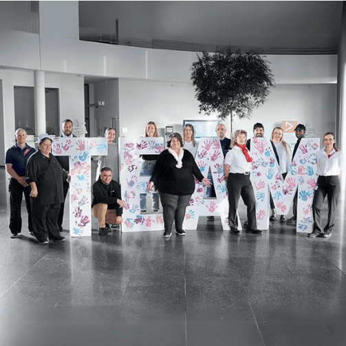 Eine Gruppe von Sodexo-Mitarbeiter:innen posiert im offenen Eingangsbereichs eines Kunden-Standorts mit großen Styropor-Buchstaben, die das Wort Team ergeben.