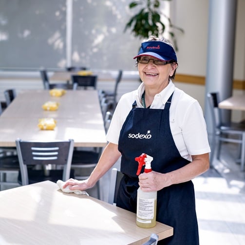 Eine Sodexo-Mitarbeiterin mit blauer Schürze und Schirmmütze reinigt die Tische in einem Betriebsrestaurant.