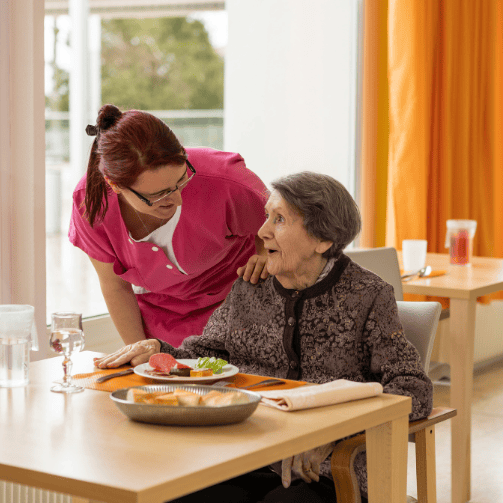 Mitarbeiterin einer Senioreneinrichtung kümmert sich freundlich um Bewohnerin, die lachend am Tisch, gedeckt mit Essen, sitzt.